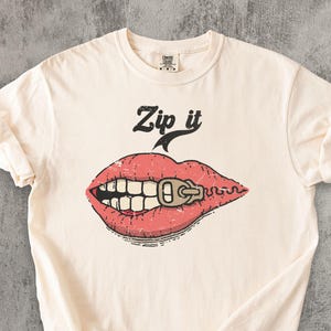 Op de afbeelding: Crèmekleurig T-shirt met een afbeelding van rode lippen met een ritssluiting en de woorden "Zip it" in het zwart. Het ontwerp heeft een vintage, distressed look. Het shirt is gemaakt van een zacht materiaal en heeft korte mouwen.