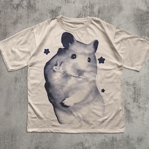 Könnte beinhalten: Cremefarbenes T-Shirt mit einem großen, stilisierten Hamster-Motiv in Marineblau mit einem gepunkteten Muster. Drei marineblaue Sternformen sind um den Hamster herum verstreut. Das T-Shirt hat kurze Ärmel und einen Rundhalsausschnitt.