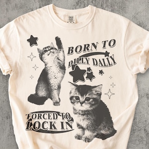 May include: Cream-coloured t-shirt with two grayscale kitten illustrations and the text "BORN TO DILLY DALLY FORCED TO LOCK IN." The design includes star graphics. The shirt is made of a soft, comfortable material.