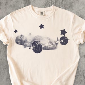 May include: Cream-colored t-shirt featuring a graphic of a person lifting a barbell, with three dark blue stars. The design is in a halftone style. The shirt has a crew neck and short sleeves.
