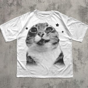 May include: White t-shirt featuring a black and white halftone image of a cat's face. The cat has large eyes and a slightly open mouth. Three black star graphics are placed around the cat's head. The shirt has short sleeves and a crew neck.