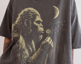 Peut inclure: Un t-shirt gris anthracite à manches courtes avec une grande impression graphique représentant une créature poilue soufflant sur un pissenlit. Le motif inclut un croissant de lune et des sapins en jaune clair sur le tissu sombre du vêtement.