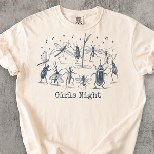 Pode incluir: Uma t-shirt de manga curta cor de creme com um gráfico em estilo de tinta azul escuro que mostra vários insetos a dançar. O design inclui notas musicais, estrelas e o texto Girls Night impresso numa fonte simples sob os insetos.