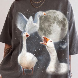 Puede incluir: Una camiseta de manga corta en color gris carbón con un estampado gráfico de tres ocas blancas en varias posturas sobre un fondo de luna llena y cielo estrellado. Las ocas presentan picos de color naranja y un plumaje detallado.