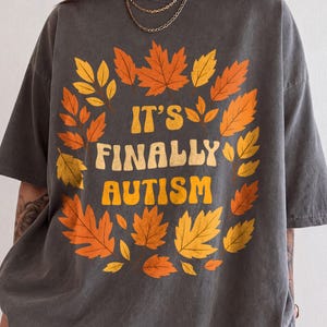 Può includere: Una t-shirt grigio antracite a maniche corte con una corona circolare di foglie autunnali arancioni, gialle e rosse che circonda il testo giallo IT'S FINALLY AUTISM in un carattere retro audace centrato sulla parte anteriore del capo.