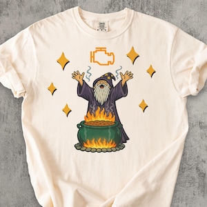 May include: Cream-coloured t-shirt with a cartoon wizard in a purple robe and hat, with a long white beard. The wizard stands before a cauldron with flames, and the check engine light symbol is above him. The design includes yellow stars.