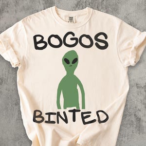 May include: Cream-colored t-shirt with the words "BOGOS BINTED" in black block letters. A green alien figure is centered below the word "BOGOS". The shirt is made of a soft material and has a casual, relaxed fit.