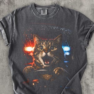 May include: Dark gray t-shirt featuring a tabby cat driving a car with emergency lights in the background. The cat has a surprised expression with its mouth open. The shirt has rolled-up sleeves.