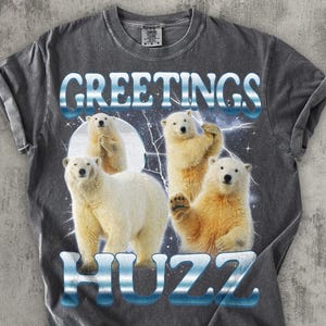 May include: Dark gray t-shirt featuring a graphic of four polar bears with the words "GREETINGS HUZZ" in a blue and white font. The bears are set against a backdrop of a moon and lightning.