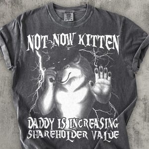 Puede incluir: Camiseta gris oscuro con un diseño de lobo y rayos. El texto en la camiseta dice "NOT NOW KITTEN" y "DADDY IS INCREASING SHAREHOLDER VALUE". Las mangas están dobladas.