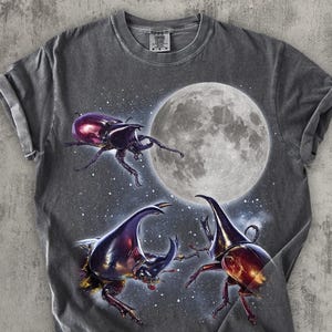 Puede incluir: Camiseta gris oscuro con un diseño de tres escarabajos rinoceronte frente a una luna llena y un fondo estrellado. Los escarabajos están representados en tonos morados, rojos y marrones.