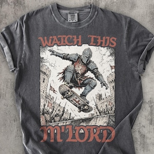 Op de afbeelding: Een antracietgrijs t-shirt met korte mouwen en een grafische print van een ridder in harnas die op een skateboard rijdt voor een kasteel. De tekst WATCH THIS M'LORD staat in een vetgedrukt rood lettertype boven en onder de illustratie.