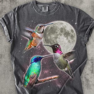 Peut inclure: T-shirt gris foncé avec une illustration colorée de trois colibris sur fond de pleine lune et de ciel étoilé. Les oiseaux présentent un plumage aux couleurs vives, avec des nuances de vert, de bleu, d'orange et de rose.