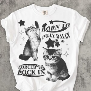 May include: White t-shirt with two grayscale cat illustrations and star graphics. The text reads "BORN TO DILLY DALLY" and "FORCED TO LOCK IN". The shirt has a crew neck and short sleeves.