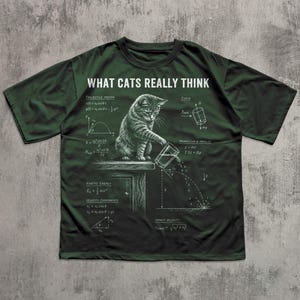 May include: A dark green t-shirt featuring a white graphic of a cat pushing a glass off a table, surrounded by physics equations and the text WHAT CATS REALLY THINK. The design includes diagrams for projectile motion, force, and kinetic energy.