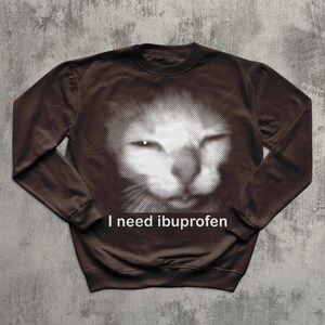 May include: Brown sweatshirt featuring a white halftone image of a cat's face. The text "I need ibuprofen" is printed in white below the cat image. The sweatshirt has long sleeves and a crew neck.