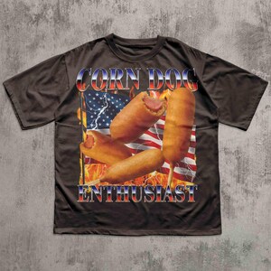 Op de afbeelding: Donkerbruin T-shirt met een afbeelding van corn dogs voor een Amerikaanse vlag met vlammen en bliksem. De tekst "CORN DOG ENTHUSIAST" is in rood, wit en blauw.