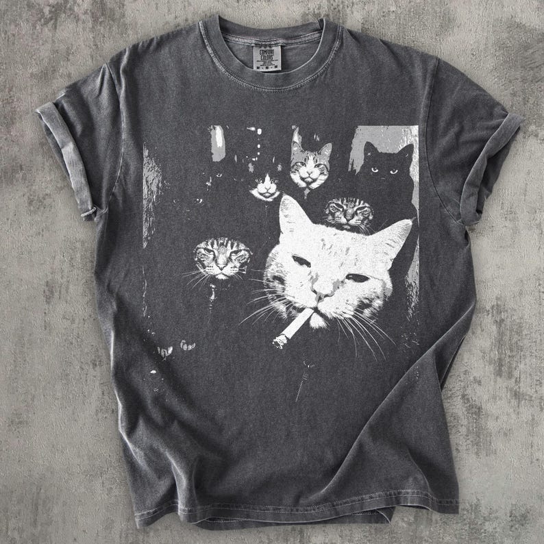 May include: Dark gray t-shirt featuring a black and white graphic of several cats, with one cat smoking a cigarette. The cats have various expressions. The shirt has rolled-up sleeves and a crew neck.