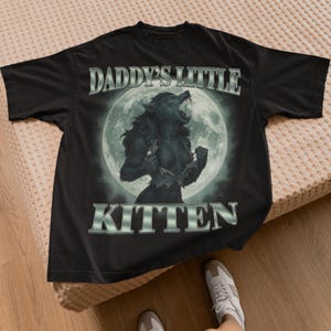 Pode incluir: Camiseta preta com uma ilustra&ccedil;&atilde;o de lobisomem em frente a uma lua cheia. O texto "DADDY'S LITTLE KITTEN" &eacute; exibido acima e abaixo da imagem em uma fonte prateada e met&aacute;lica. A camiseta &eacute; feita de um material macio.