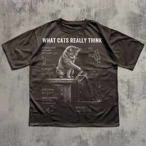 May include: A dark brown t-shirt featuring a white graphic of a cat pushing a glass off a table, surrounded by physics equations and diagrams. The text "WHAT CATS REALLY THINK" is printed in white at the top of the shirt design.