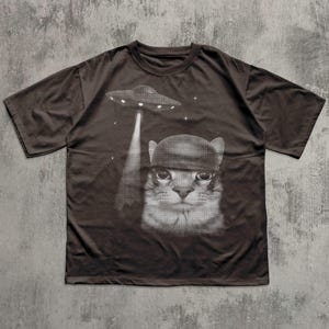 K&ouml;nnte beinhalten: Dunkelbraunes T-Shirt mit einer schwarz-wei&szlig;en Grafik einer Katze mit Hut, die von einem UFO entf&uuml;hrt wird. Das Design beinhaltet Sterne. Das T-Shirt hat einen Rundhalsausschnitt und kurze &Auml;rmel.