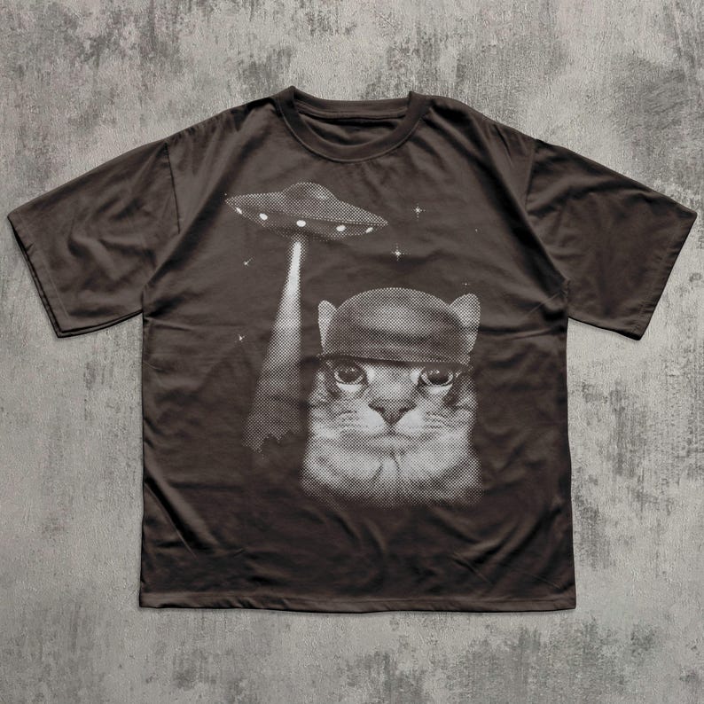 K&ouml;nnte beinhalten: Dunkelbraunes T-Shirt mit einer schwarz-wei&szlig;en Grafik einer Katze mit Hut, die von einem UFO entf&uuml;hrt wird. Das Design beinhaltet Sterne. Das T-Shirt hat einen Rundhalsausschnitt und kurze &Auml;rmel.