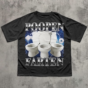 Op de afbeelding: Zwart T-shirt met een grafisch ontwerp met drie witte toiletten, bliksemflitsen en de woorden "POOPEN FARTEN" in een zilverkleurig, chroomachtig lettertype. Een rol toiletpapier is ook zichtbaar.