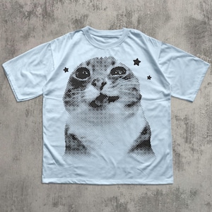 May include: Light blue t-shirt featuring a black and white halftone image of a cat's face. Three black star graphics are placed around the cat image. The shirt has short sleeves and a crew neck.