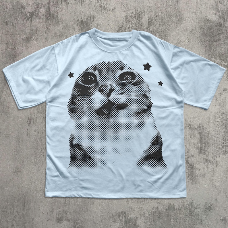 May include: Light blue t-shirt featuring a black and white halftone image of a cat's face. Three black star graphics are placed around the cat image. The shirt has short sleeves and a crew neck.