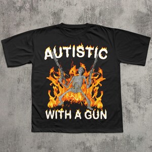 May include: Black t-shirt featuring a skeleton holding two guns, surrounded by flames. The words "AUTISTIC" and "WITH A GUN" are printed in white. The design is centered on the shirt.