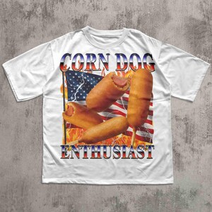 Op de afbeelding: Wit T-shirt met een afbeelding van corn dogs voor een Amerikaanse vlag met de tekst "CORN DOG ENTHUSIAST" in rood, wit en blauw. De afbeelding bevat vlammen en bliksem.