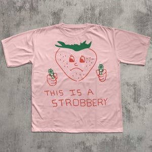 May include: A light pink t-shirt featuring a cartoon strawberry with a sad face holding two guns. The text "THIS IS A STROBBERY" is printed in red below the strawberry graphic.