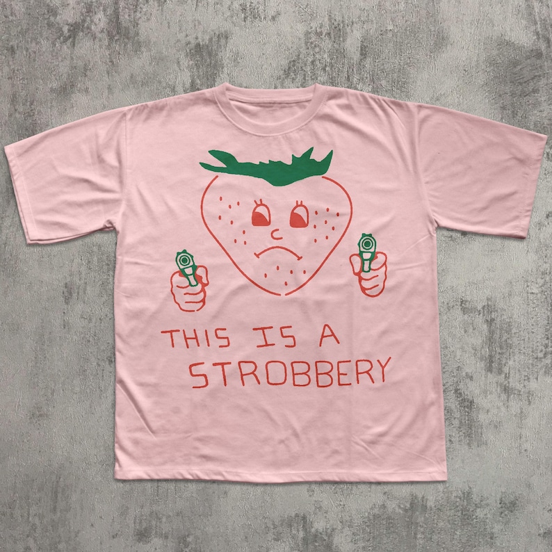 May include: A light pink t-shirt featuring a cartoon strawberry with a sad face holding two guns. The text "THIS IS A STROBBERY" is printed in red below the strawberry graphic.