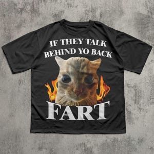 Op de afbeelding: Zwart t-shirt met een kattenafbeelding omringd door vlammen. De tekst boven de kat luidt "IF THEY TALK BEHIND YO BACK" en de tekst eronder luidt "FART" in witte letters.