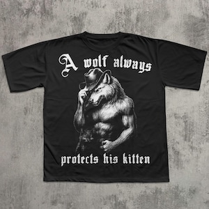 May include: Black t-shirt featuring a detailed illustration of a wolf wearing a hat, with the text "A wolf always protects his kitten" in white gothic lettering. The wolf is depicted with a muscular physique.