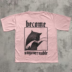 Op de afbeelding: Lichtroze T-shirt met een zwarte afbeelding van een kat die in een hand bijt, omlijst door een zwart rechthoek. De woorden "become" en "ungovernable" staan in gotische letters boven en onder de afbeelding.