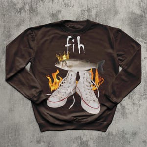 May include: A brown sweatshirt with a fish wearing a gold crown above white high-top trainers. The word "fih" is written above the fish in white. Flames are visible behind the trainers.