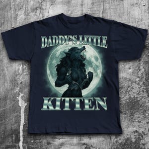 Pode incluir: Camiseta azul marinho com um gr&aacute;fico de lobisomem em frente a uma lua cheia. O texto "DADDY'S LITTLE KITTEN" &eacute; exibido acima e abaixo da imagem em uma fonte met&aacute;lica prateada. O lobisomem est&aacute; em transforma&ccedil;&atilde;o.