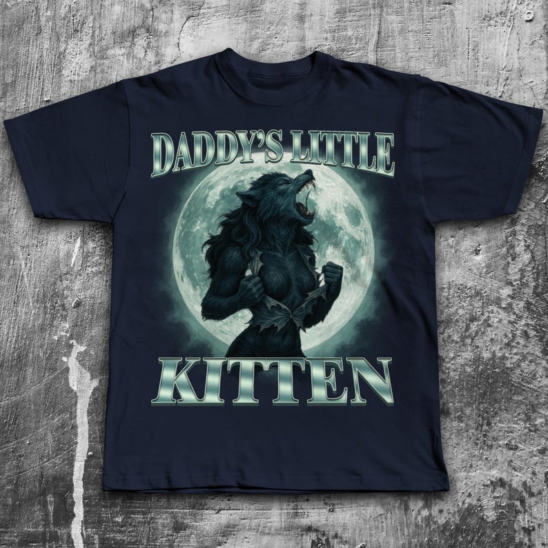 Pode incluir: Camiseta azul marinho com um gr&aacute;fico de lobisomem em frente a uma lua cheia. O texto "DADDY'S LITTLE KITTEN" &eacute; exibido acima e abaixo da imagem em uma fonte met&aacute;lica prateada. O lobisomem est&aacute; em transforma&ccedil;&atilde;o.