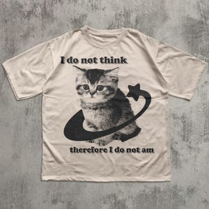 May include: Cream-colored t-shirt featuring a black and white illustration of a kitten encircled by a curved line and star. The text "I do not think therefore I do not am" is printed above and below the cat graphic. The shirt has short sleeves.