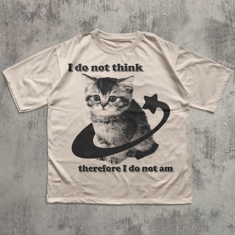 May include: Cream-colored t-shirt featuring a black and white illustration of a kitten encircled by a curved line and star. The text "I do not think therefore I do not am" is printed above and below the cat graphic. The shirt has short sleeves.