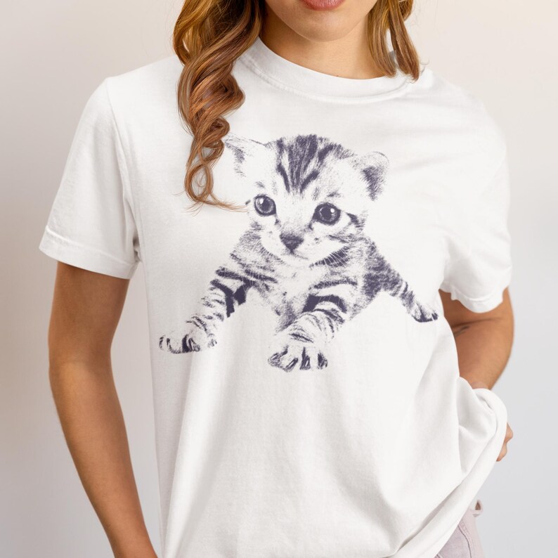 K&ouml;nnte beinhalten: Wei&szlig;es T-Shirt mit einer schwarz-wei&szlig;en Illustration eines K&auml;tzchens. Das K&auml;tzchen hat gro&szlig;e Augen und gestreifte Markierungen. Das T-Shirt hat einen klassischen Rundhalsausschnitt.