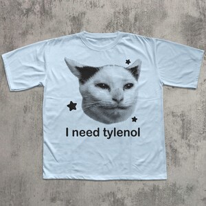 May include: Light blue t-shirt featuring a black and white cat face graphic with three black stars. The text "I need tylenol" is printed below the cat face. The shirt has short sleeves and a crew neck.