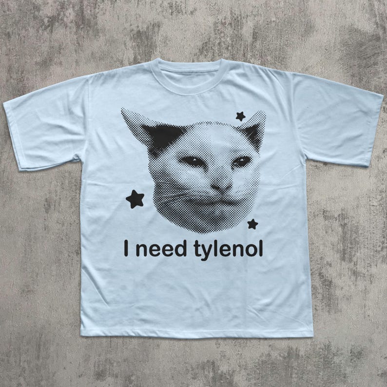 May include: Light blue t-shirt featuring a black and white cat face graphic with three black stars. The text "I need tylenol" is printed below the cat face. The shirt has short sleeves and a crew neck.