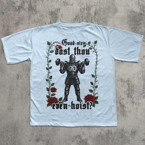 May include: Light blue t-shirt featuring a medieval knight lifting dumbbells, framed by roses and vines. The text reads "Good sir dost thou even hoist?" in a gothic font. A unique design for fitness enthusiasts.