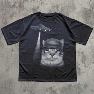 K&ouml;nnte beinhalten: Dunkelblaues T-Shirt mit einem schwarz-wei&szlig;en Grafikdesign einer Katze mit Helm, die von einem UFO entf&uuml;hrt wird. Das Design enth&auml;lt Sterne. Das T-Shirt hat kurze &Auml;rmel und einen Rundhalsausschnitt.