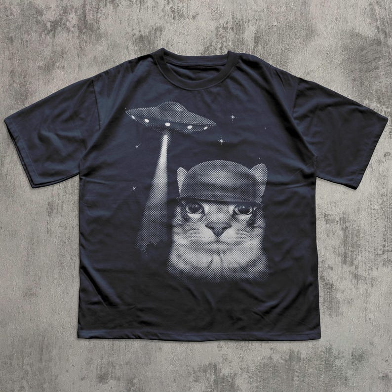 K&ouml;nnte beinhalten: Dunkelblaues T-Shirt mit einem schwarz-wei&szlig;en Grafikdesign einer Katze mit Helm, die von einem UFO entf&uuml;hrt wird. Das Design enth&auml;lt Sterne. Das T-Shirt hat kurze &Auml;rmel und einen Rundhalsausschnitt.