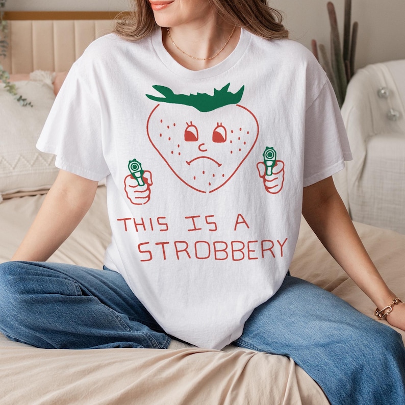 May include: White t-shirt with a red strawberry graphic holding two green toy guns. The strawberry has a sad face and the text "THIS IS A STROBBERY" is printed below the graphic.