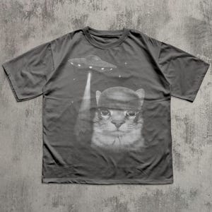 K&ouml;nnte beinhalten: Dunkelgraues T-Shirt mit einem schwarz-wei&szlig;en Grafikdesign einer Katze mit Hut, die von einem UFO entf&uuml;hrt wird. Das Design enth&auml;lt Sterne. Das T-Shirt ist ein l&auml;ssiger Stil mit kurzen &Auml;rmeln.