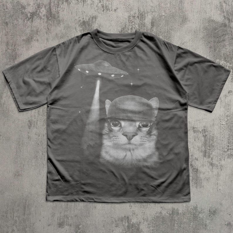 K&ouml;nnte beinhalten: Dunkelgraues T-Shirt mit einem schwarz-wei&szlig;en Grafikdesign einer Katze mit Hut, die von einem UFO entf&uuml;hrt wird. Das Design enth&auml;lt Sterne. Das T-Shirt ist ein l&auml;ssiger Stil mit kurzen &Auml;rmeln.